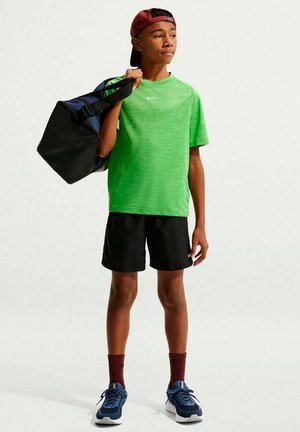 Jongen in een groen sportshirt, zwarte korte broek, rode pet en blauwe sneakers draagt een zwarte en blauwe sporttas over één schouder.