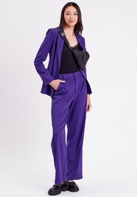 Costume ajusté violet avec un pantalon à jambes larges, tissu texturé, et un sac bandoulière matelassé noir sur un haut noir, porté avec des chaussures imposantes.