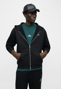 Νεαρός άνδρας που φοράει μαύρη ζακέτα με φερμουάρ, πράσινο μπλουζάκι με λογότυπο, μαύρη φόρμα και μαύρο δερμάτινο καπέλο μπέιζμπολ, με τα χέρια στις τσέπες της ζακέτας.