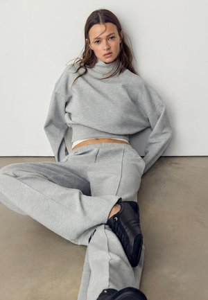 Giovane donna con capelli castani lunghi che indossa una felpa e pantaloni grigi, seduta sul pavimento con scarpe da ginnastica nere contro una parete chiara.