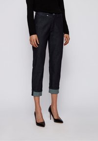 Jeans oscuros de mezclilla con puños enrollados que presentan un patrón geométrico en contraste. Corte ajustado con bolsillos frontales y detalles de hardware metálico.