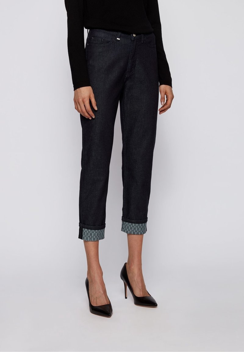 Jeans oscuros de mezclilla con puños enrollados que presentan un patrón geométrico en contraste. Corte ajustado con bolsillos frontales y detalles de hardware metálico.