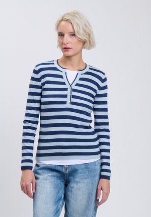 Vrouw met kort blond haar die een blauw en lichtblauw gestreept shirt met lange mouwen en lichtblauwe spijkerbroek draagt tegen een effen achtergrond.