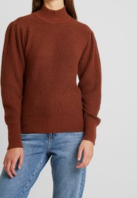 Bruine geribbelde trui met een hoge hals en een gestructureerd patroon; heeft opgeblazen mouwen en een aansluitende tailleband, gecombineerd met lichtblauwe spijkerbroek.