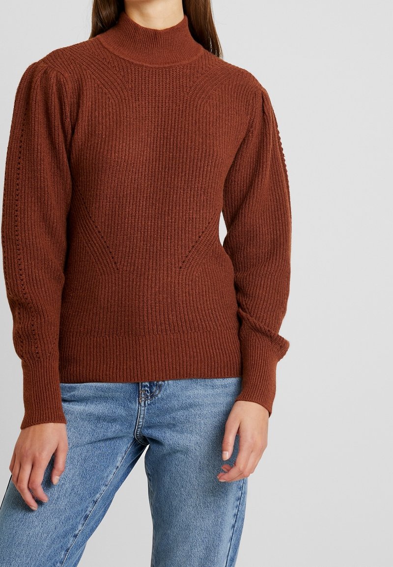 Bruine geribbelde trui met een hoge hals en een gestructureerd patroon; heeft opgeblazen mouwen en een aansluitende tailleband, gecombineerd met lichtblauwe spijkerbroek.