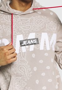 Felpa beige chiaro con motivo paisley e lettering bianco prominente che recita "JEANS". Presenta un cappuccio con cordino rosso e tasca frontale.