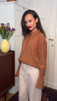 Γυναίκα με μακριά σκούρα μαλλιά, φορώντας μια μπλούζα με χρώμα σκουριάς και κρεμ παντελόνι, στέκεται κοντά σε ένα ντουλάπι με ένα κίτρινο ριγέ βάζο γεμάτο μωβ και λευκά λουλούδια.