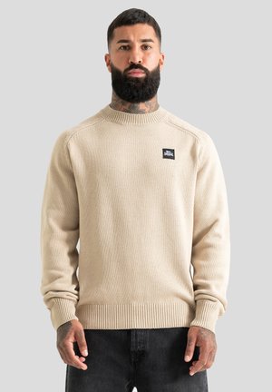 Homme avec barbe et tatouages portant un pull en maille beige et un pantalon noir, debout devant un fond gris clair uni.