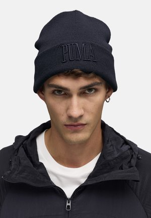 Young man wearing a black Puma beanie, black jacket with a zipper, white shirt underneath, and a small hoop earring in one ear.