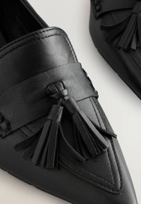 Mocassins en cuir noir avec bout pointu, pompons décoratifs et une large bride sur le dessus. Texture lisse avec des détails cousus.