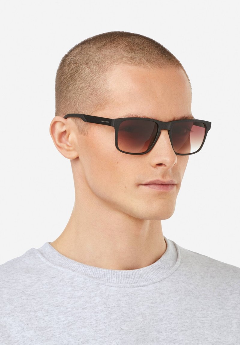 Mustat suorakaiteen muotoiset aurinkolasit, joissa on liukuväri ruskeat linssit. Lasit ovat mattapintaiset ja minimalistisen tyylikkäät.