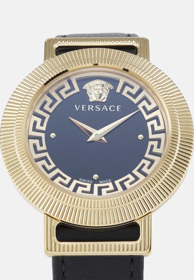 Reloj Versace en tono dorado con esfera azul marino, intrincado patrón de clave griega, emblema de león y correa de cuero negra. Fabricado en Suiza.