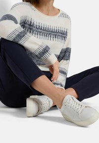 En person som sitter i korslagda ben, iförd en vit tröja med marinblå mönster, marinblå byxor och vita snörskor, på en vit bakgrund.