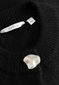 Černý pletený svetr s žebrovanou texturou a bílou broží ve tvaru abstraktního motivu. Štítky uvádějí značku a původ.