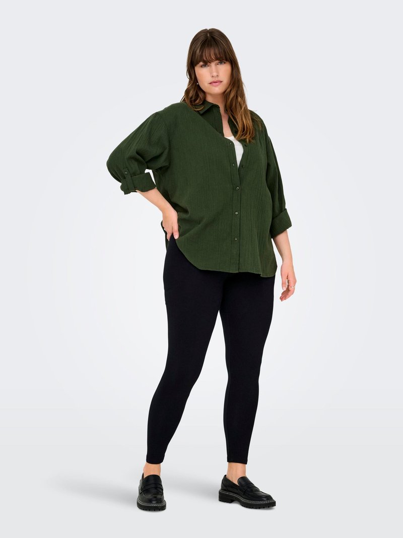 Camicia button-up oversize di colore verde scuro con maniche arrotolate, canotta bianca sotto, leggings neri e scarpe slip-on nere. Texture liscia.