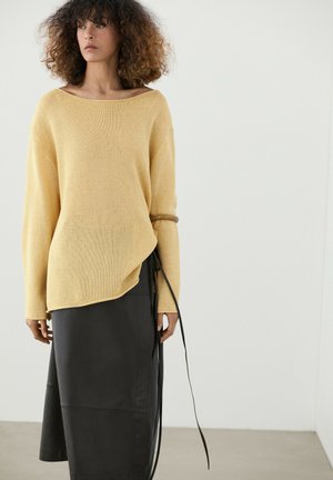 Kvinde med krøllet hår iført en løs gul sweater og sort midi nederdel med lange sorte bånd mod en ensfarvet hvid baggrund.
