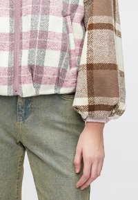 Mitmevärviline ruuduline jakk roosade, hallide ja pruunide toonidega; varustatud tõmbluku ja elastsete varrukasäärtega. Kantud heledate teksapükste peal.