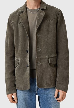Mann trägt olivgrüne Wildlederjacke über passendem Pullover und blauen Jeans, mit sichtbarer silberner Kettenkette um den Hals.