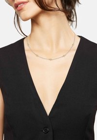 Collar de plata con acentos dorados, que presenta pequeños eslabones circulares espaciados uniformemente. Llevado sobre una blusa negra de escote en V, mostrando un diseño delicado.