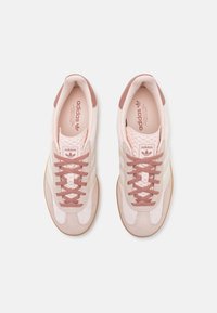 Par rosa Adidas-sneakers med bruna skosnören, beige ränder och sulor av gummi, sedd ovanifrån på vit bakgrund.