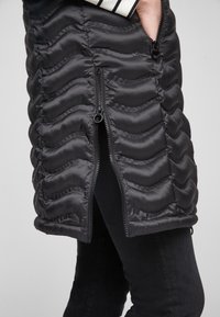 Schwarze Steppweste mit einem gewellten, gesteppten Design und zwei seitlichen Reißverschlüssen. Das Material ist glatt und leicht glänzend, was für Textur sorgt.