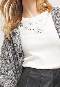 Cardigan en maille grise avec boutons noirs, porté sur un pull blanc orné d'un motif floral en perles au niveau de l'encolure, assorti à une jupe noire.