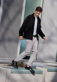 Un homme descendant d'un rebord, portant une veste noire, une chemise blanche, un pantalon gris clair et des baskets noires, à l'extérieur près d'un bâtiment moderne.