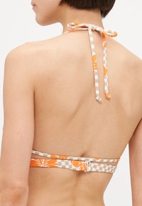 Bikini zgoraj z halter vratom, ki ima oranžno cvetlično oblikovanje na bež podlagi s karo detajli. Zavezane naramnice na hrbtu omogočajo prilagodljivo prileganje.
