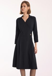 Czarna sukienka kopertowa z długimi rękawami, kołnierzykiem, plisowanym dołem i guzikiem w talii. Gładki materiał i dopasowany krój.