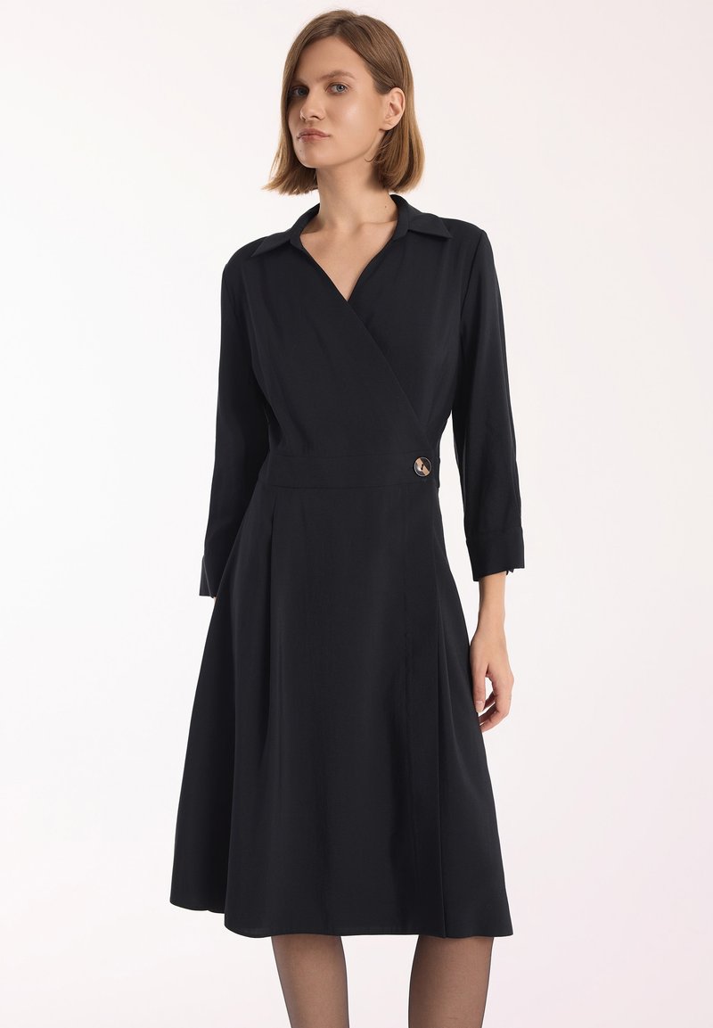 Czarna sukienka kopertowa z długimi rękawami, kołnierzykiem, plisowanym dołem i guzikiem w talii. Gładki materiał i dopasowany krój.