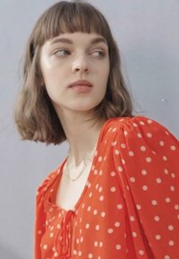 Blusa vermelha com bolinhas, com mangas bufantes, apresentando um laço na gola. A modelo tem cabelo até os ombros e um fundo neutro.