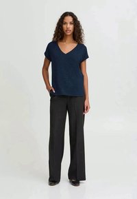 Woman with curly hair wearing a navy blue short-sleeve V-neck top and black wide-leg trousers, standing with one hand in pocket.