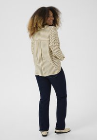 Blusa a rayas en beige y blanco con un corte holgado, combinada con jeans azul oscuro y zapatos de color claro, vista desde la parte de atrás.