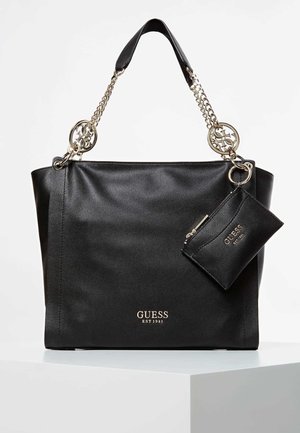 Sac fourre-tout noir Guess avec anses à chaînes dorées, petite pochette assortie attachée, et logo Guess en lettres dorées sur le devant.