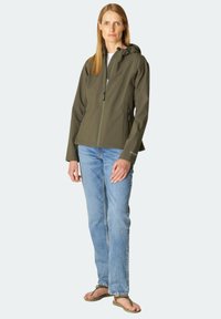 Veste imperméable vert olive avec fermeture éclair sur le devant, capuche ajustable et deux poches latérales, associée à un jean bleu clair et des tongs.