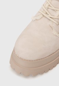 Detailní záběr na béžovou semišovou botu s tlustou vroubkovanou podrážkou a huňatým lemováním kolem očí pro tkaničky na světlém pozadí.