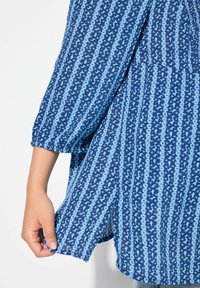Blaue Bluse aus sternenmusterndem Stoff, mit lockerer Passform, gerafften 3/4-Ärmeln und abgerundetem Saum. Das Material wirkt weich und leicht.
