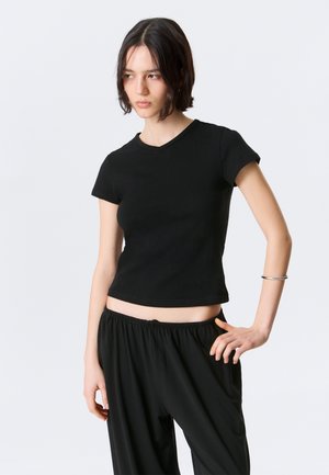 Jeune femme aux cheveux courts noirs, portant un haut noir à manches courtes et un pantalon noir ample, main sur la hanche, sur un fond uni.