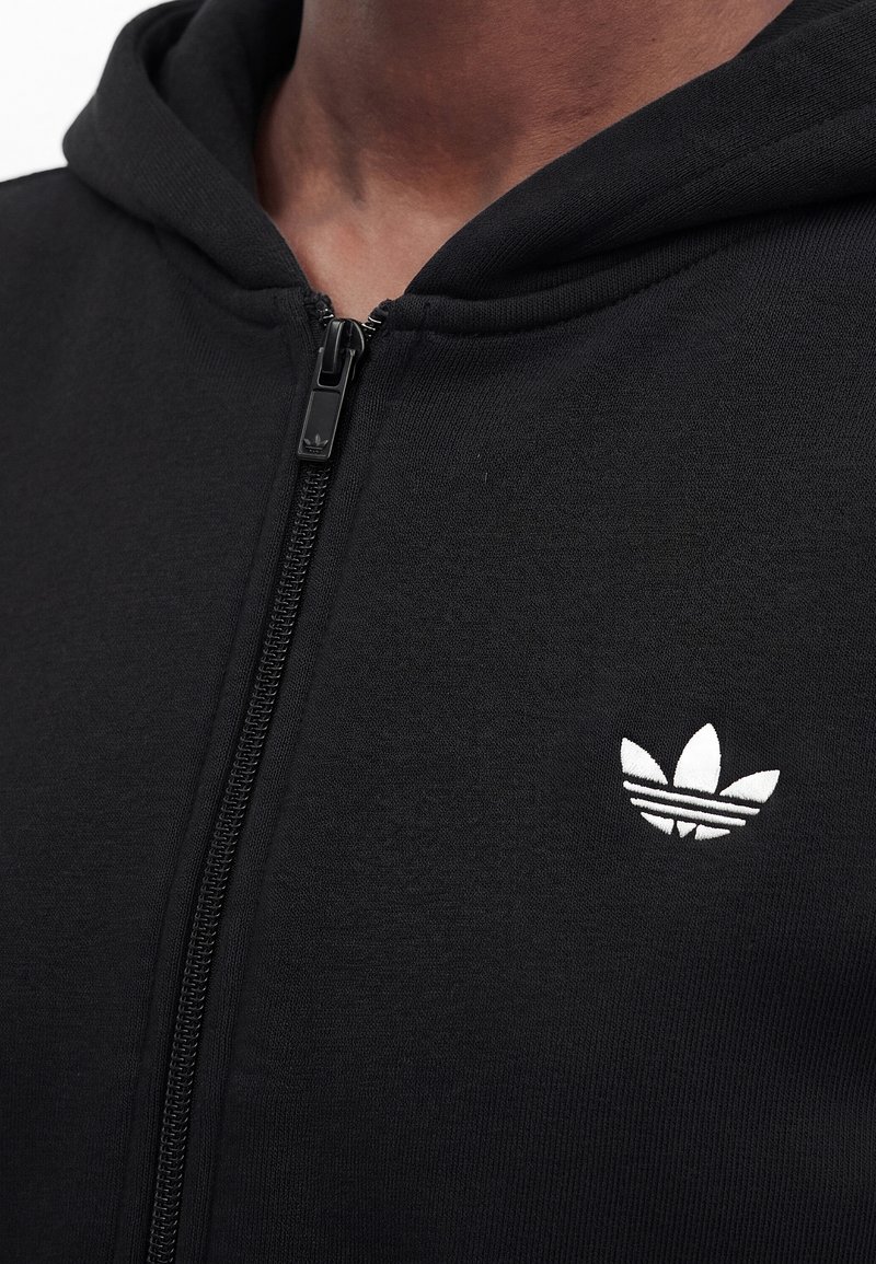 Gros plan d'une personne portant un sweat à capuche noir zippé avec un logo Adidas Trefoil blanc sur la poitrine.