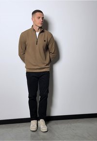 Hnědý svetr na zip s černým akcentem, kombinovaný s černými kalhotami a krémovými teniskami. Model stojí na jednoduchém bílém pozadí.