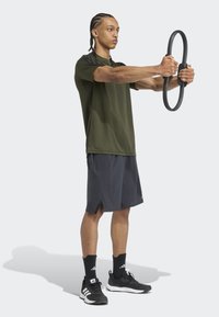 Olivovo zelené športové tričko s čiernymi akcentmi, šedé šortky, drží kruhový čierny cvičebný nástroj, čierne športové topánky, stojí s vzpriameným postojom.