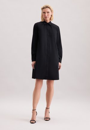 Femme debout portant une robe chemise noire à manches longues et des sandales à talons hauts noires avec bride à la cheville sur un fond uni.