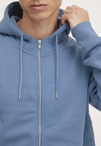 Sudadera con cremallera azul claro, hecha de un tejido suave, que cuenta con capucha con cordón y cierre de cremallera metálico. Textura suave con puños acanalados.