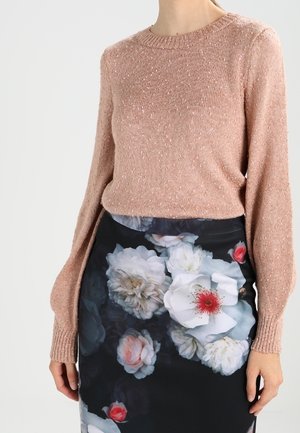 Kobieta ubrana w teksturowany sweter w kolorze przydymionego różu, w połączeniu z czarną spódnicą z dużymi białymi i różowymi kwiatowymi wzorami.