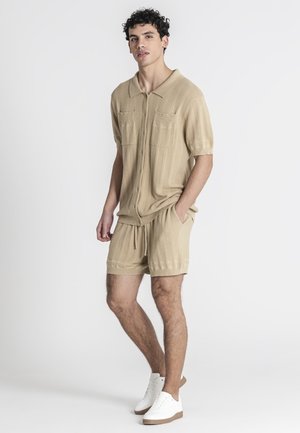 Hombre está de pie con las manos en los bolsillos, vistiendo una camisa de punto beige de manga corta con botones y pantalones cortos a juego con zapatillas blancas sobre fondo blanco.
