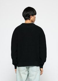 Personen vender bort med en svart strikkegenser og lyseblå jeans, stående mot en enkel hvit bakgrunn.