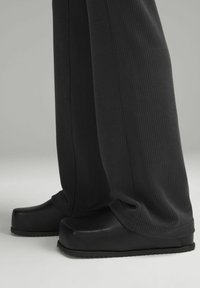 Musta puukengät, joilla on sileä, kiiltävä pinta ja tukeva muoto sekä uritettujen pohjien varusteena, yhdistettyinä löysiin, ribattuihin mustiin housuihin.