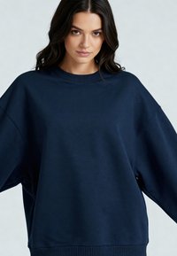 Mornarsko modra oversized majica s kapuco, izdelana iz mehkega materiala, z okroglim ovratnikom, širokimi rokavi ter rebrastimi manšetami in robom.