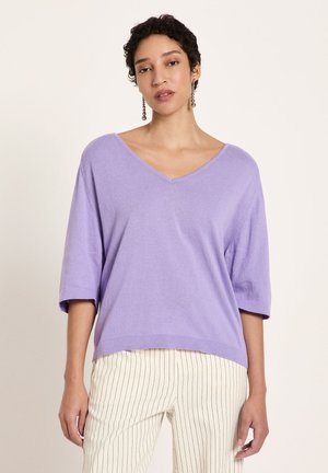 Frau trägt einen lockeren lavendelfarbenen V-Ausschnitt-Pullover mit ellbogenlangen Ärmeln und weiße Hosen mit dünnen schwarzen Vertikalstreifen.