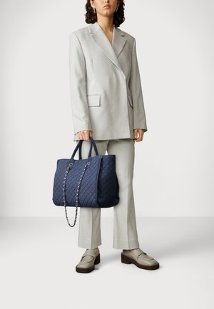 Dunkelblaues, gestepptes Shopper-Tasche mit Kettelementen, stabilen Griffen und geräumigem Design. Kombiniert mit einem hellgrauen Anzug und beigen Schuhen.